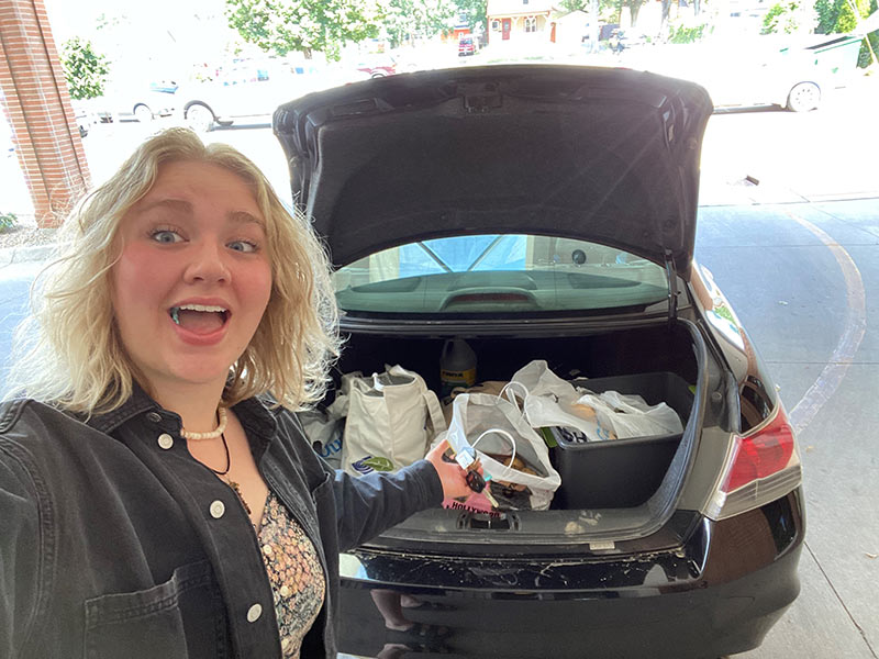 smiling woman in front of open car trunk filled with donated food